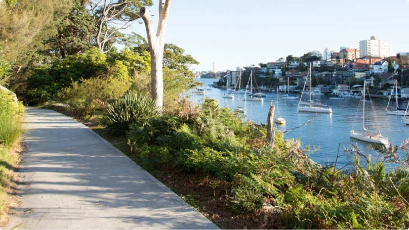 A paved path runs alongside a river with moored sailboats, leafy trees, and residential buildings visible in the background.