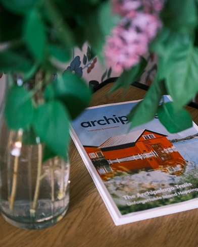 A magazine titled "archipelago" with a cover photo of a red house sits on a wooden table, partially obscured by a vase of green leaves and purple flowers.