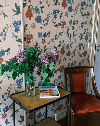 A small table with a vase of purple flowers and a magazine beside a wooden chair, set against a floral wallpapered wall.