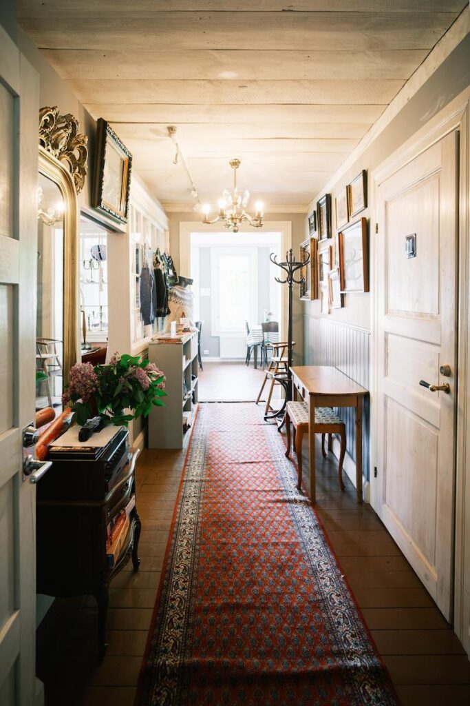 Lanternas hall med en röd mönstrad löpare, trägolv, inramade tavlor på väggarna, en spegel och en dörr som leder in till ett ljust rum i bakgrunden.