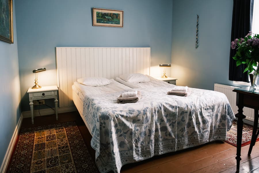 Ett välordnat sovrum med två nattduksbord, lampor, en dubbelsäng med ett mönstrat överkast, två handdukar, inramade tavlor på de blå väggarna och ett litet bord med blommor vid fönstret.