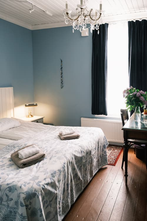 En snyggt bäddad säng med två handdukar ovanpå, en sänglampa, en ljuskrona, blå väggar och ett fönster med mörka gardiner i ett mysigt sovrum.