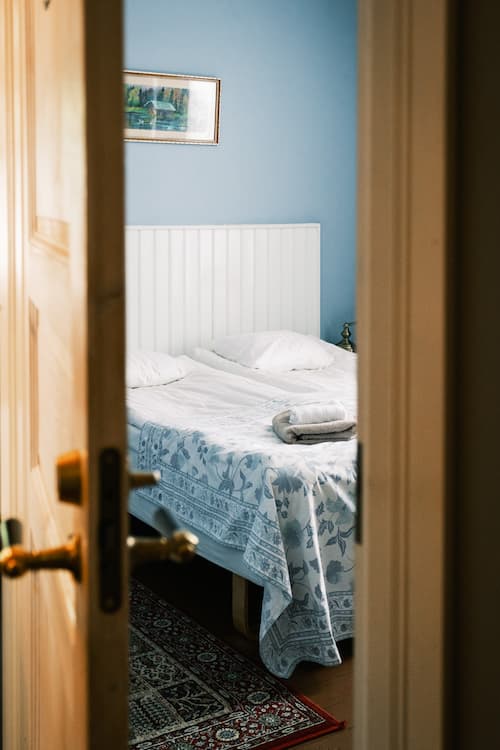Genom en halvöppen dörr syns en snyggt bäddad säng med vitt och blått sänglinne i ett svagt upplyst sovrum med blå väggar och en inramad tavla ovanför sängen.