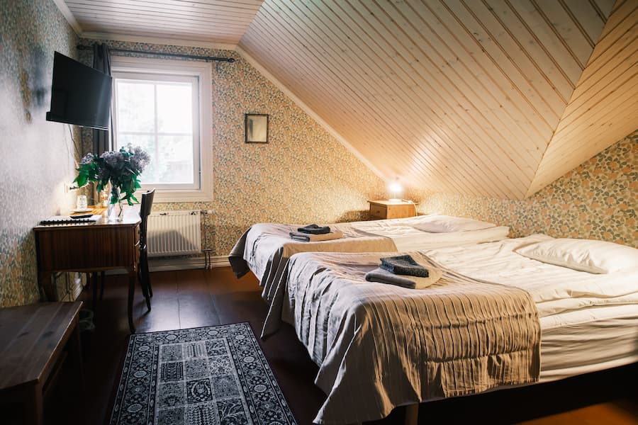 Sovrum på vinden med två enkelsängar, en TV, blommig tapet, ett litet skrivbord med blommor, ett fönster och en matta på golvet. Handdukar ligger på varje säng.