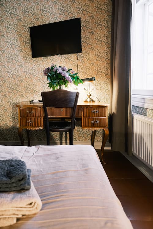 Ett skrivbord i trä med en stol, blommor och en lampa står mot en blommig tapet. Ovanför hänger en TV, och i förgrunden, nära ett fönster, står en säng med hopvikta handdukar.