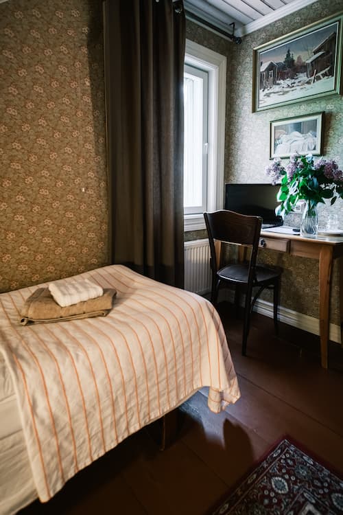 Ett litet sovrum med en enkelsäng, hopvikta handdukar, ett skrivbord med en stol, en vas med blommor, inramade bilder på väggen och ett fönster med gardiner.