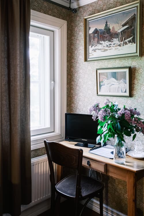 Ett litet skrivbord i trä med en datorskärm, en vas med lila blommor, en stol och en inramad tavla på en vägg med blommig tapet, intill ett fönster med bruna gardiner.