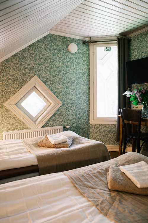 Ett mysigt sovrum på vinden med två enkelsängar, blommig tapet, ett rombformat fönster och ett skrivbord med blommor och en TV. Handdukarna ligger prydligt upplagda på varje säng.