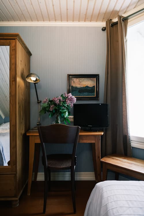 Ett litet skrivbord med en stol, en datorskärm och en vas med blommor står intill en blå vägg under ett fönster, med en garderob till vänster och en tavla ovanför skrivbordet.