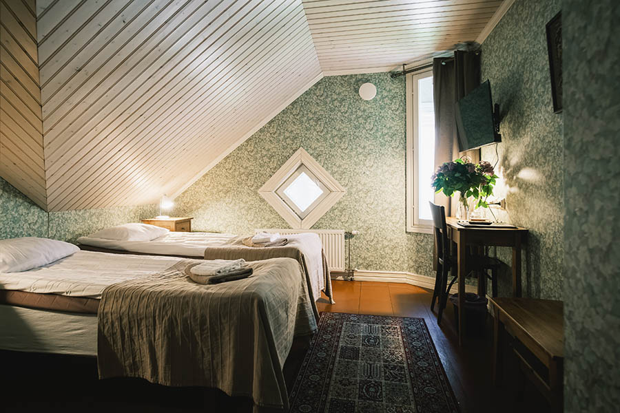 Sovrum på vinden med två enkelsängar, blommig tapet, ett litet skrivbord med en vas med blommor, en väggmonterad TV och ett fönster.
