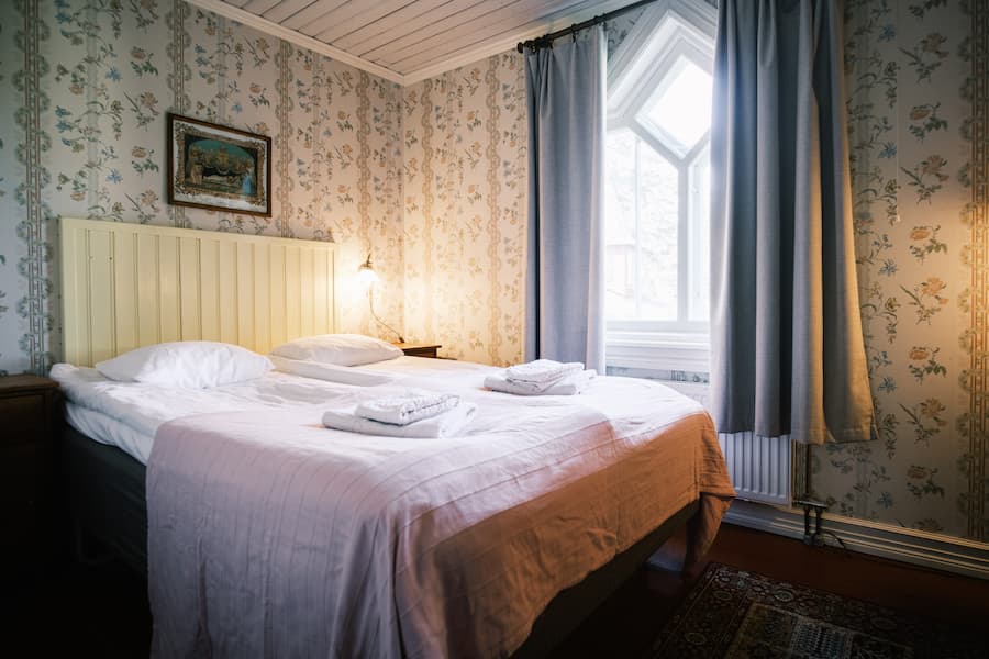 En snyggt bäddad dubbelsäng med hopvikta handdukar står i ett mysigt sovrum med blommig tapet, ett fönster med gardiner och en inramad tavla på väggen.