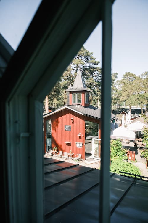 Utsikt genom ett fönster i en röd träbyggnad med spetsigt tak, omgiven av träd och uteservering i en solig miljö.
