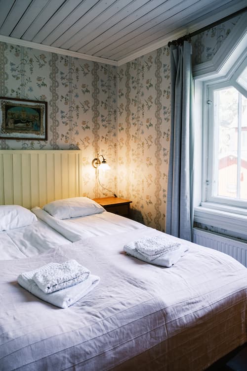 En snyggt bäddad dubbelsäng med två handdukar ovanpå, bredvid ett fönster med gardiner i ett rum med blommig tapet och en vägglampa.