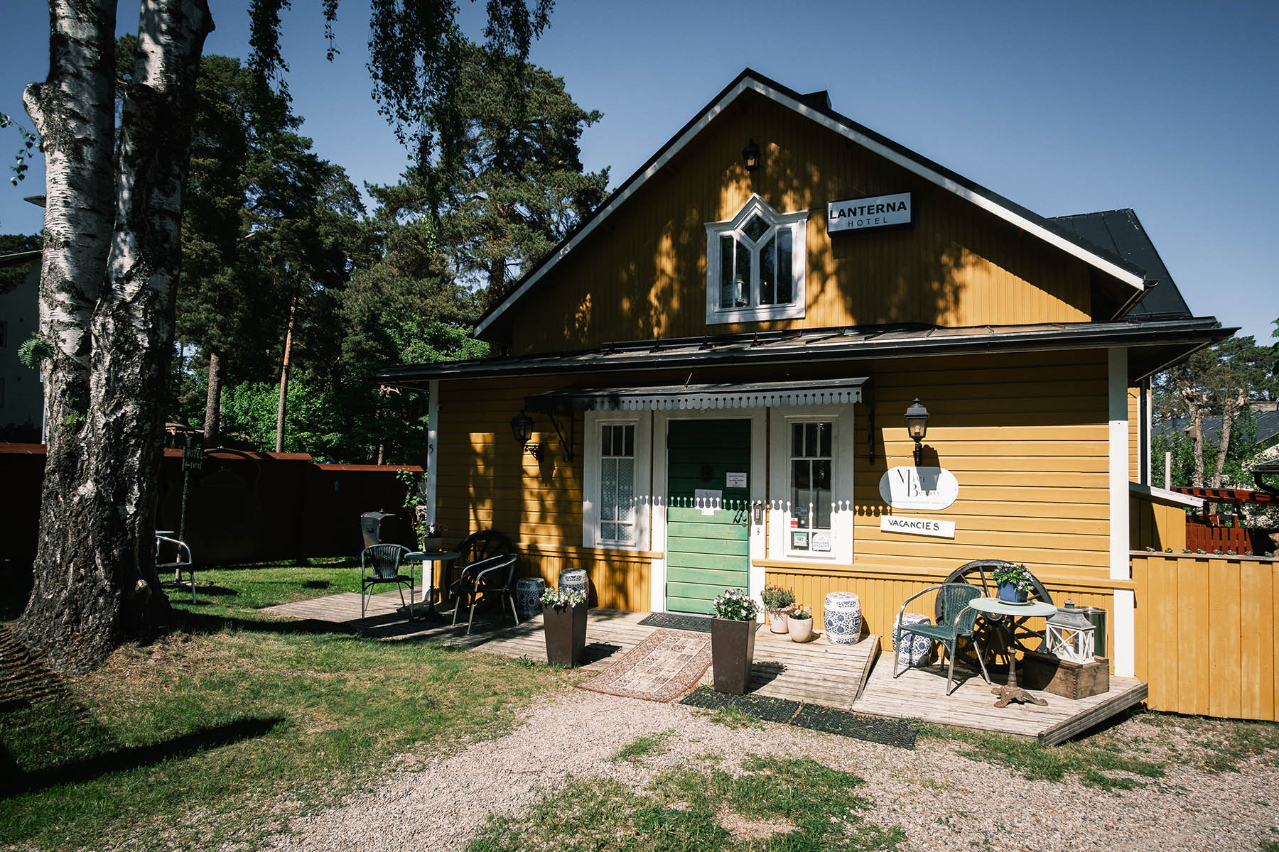 Ett gult trähus med en grön dörr, uteservering och träd i bakgrunden en solig dag.