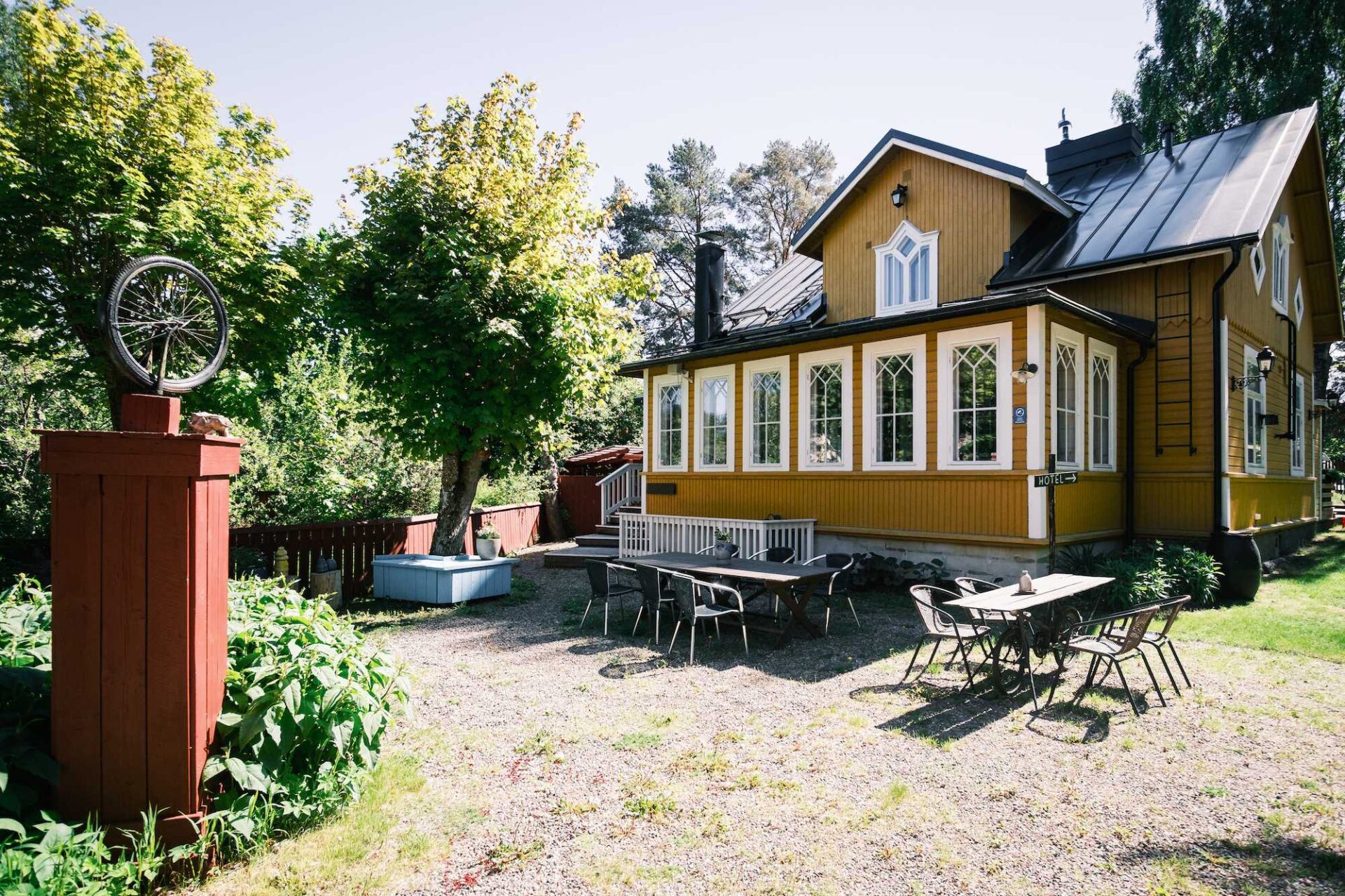 Ett gult trähus med vita lister, omgivet av träd. Utomhusbord och stolar står uppställda på grus, och i förgrunden står en röd stolpe med en skulptur föreställande ett cykelhjul.