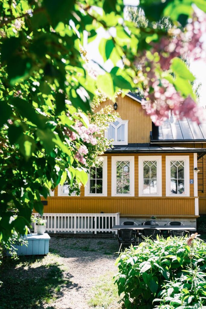 Hotel Lanterna skyms delvis av grönt lövverk och rosa blommor i en solbelyst trädgård.