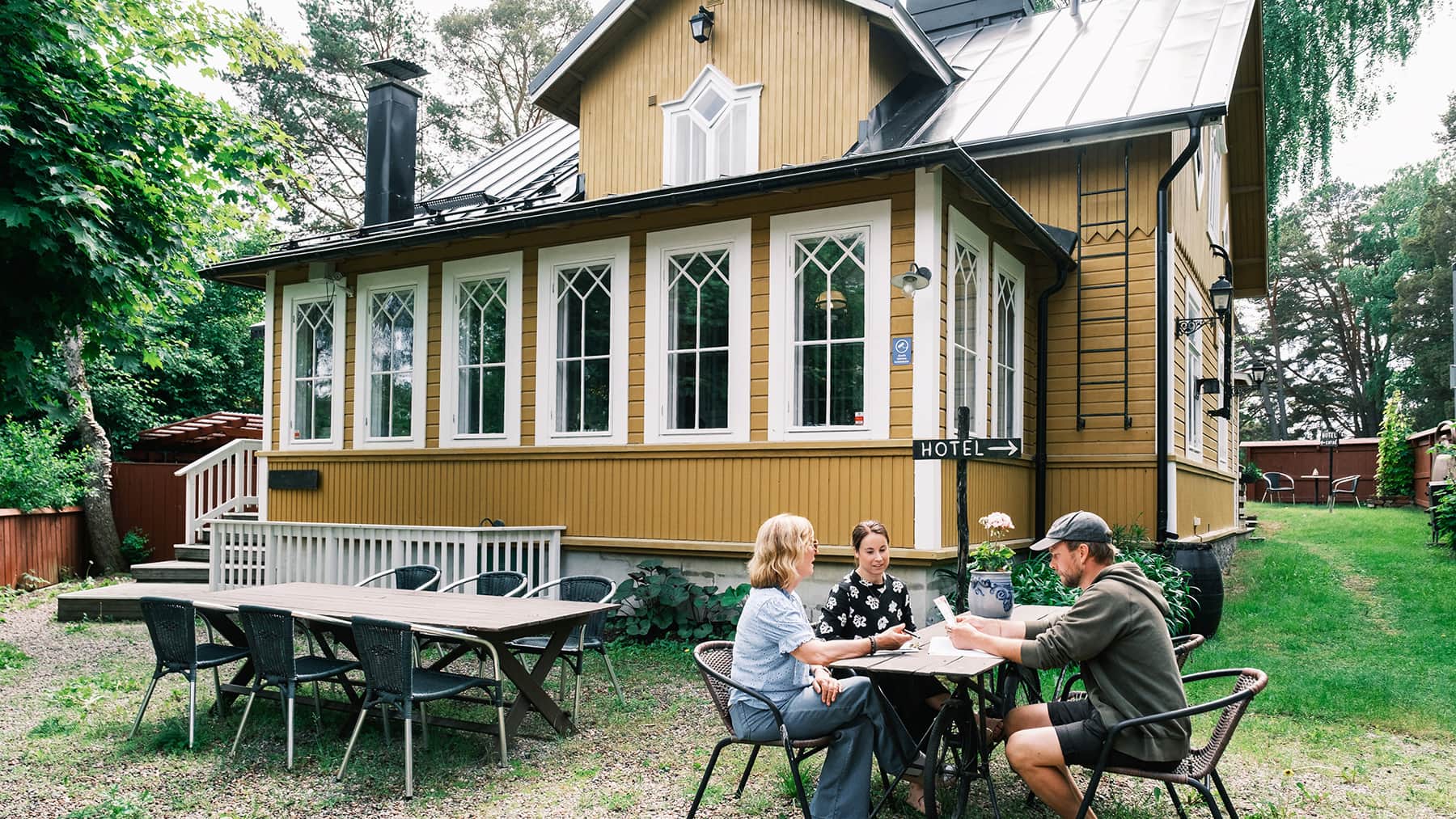 Tre personer sitter vid ett bord utanför ett gult trähus med en skylt där det står ”hotell”. I närheten finns fler tomma stolar och ett långt bord. Miljön är grön och skuggig.