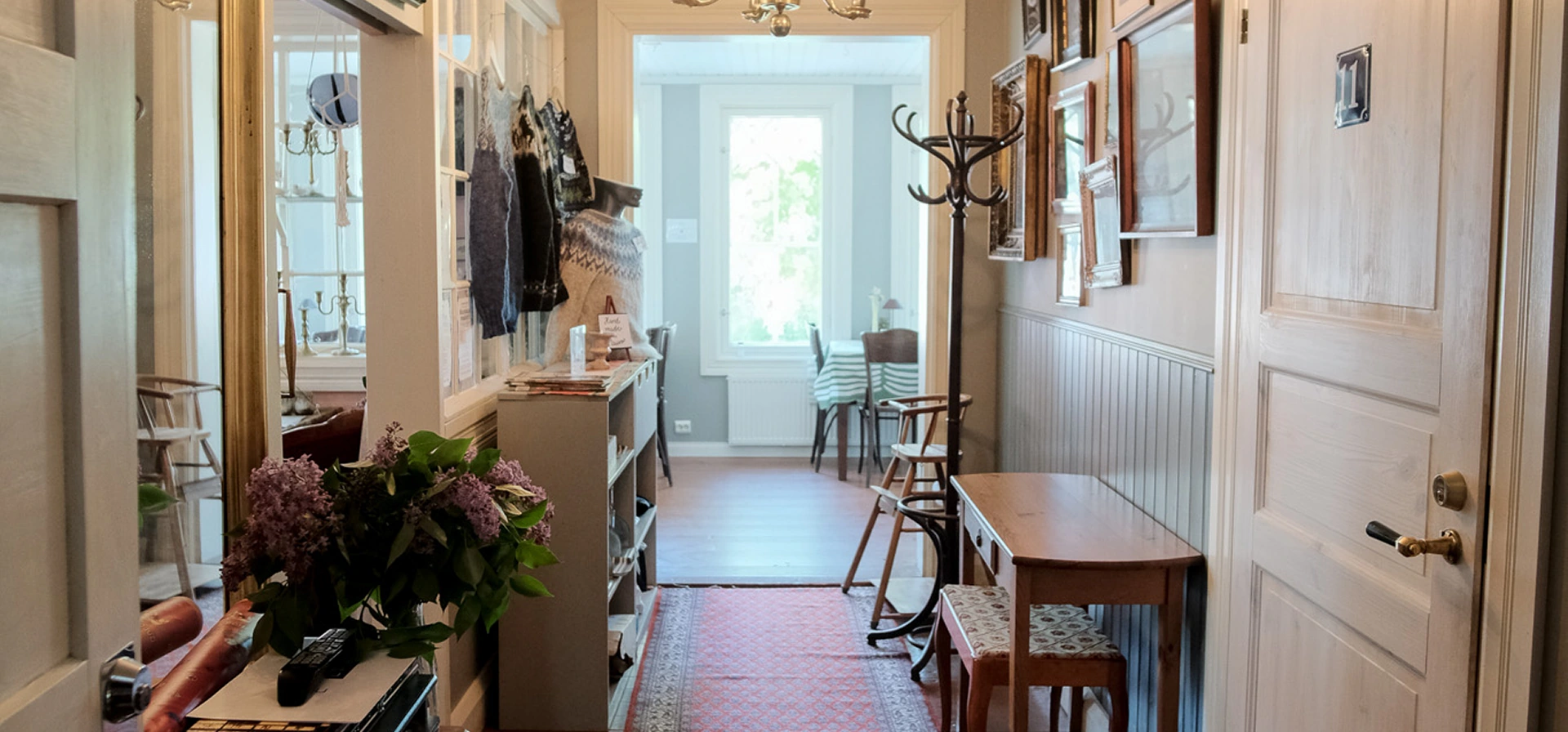 A corridor with wooden floors and light-coloured walls, decorated with framed pictures, a coat stand, a small table, and chairs, leading to a sunlit room with a window.