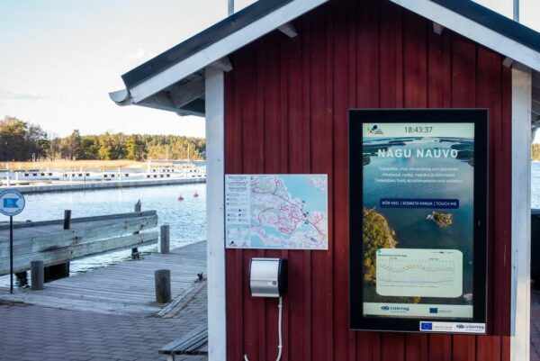 En röd träbyggnad vid en brygga är utrustad med en karta, en digital skärm med information om Nagu och en dispenser för handdesinfektionsmedel. I bakgrunden syns båtar och vatten.