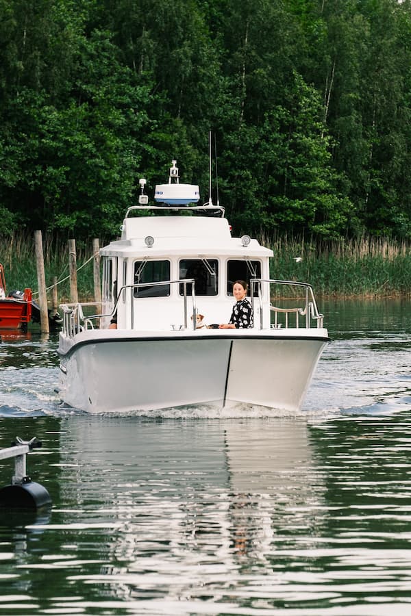 En vit motorbåt med en person vid rodret glider fram över ett lugnt vatten, med träd och vass synliga i bakgrunden.