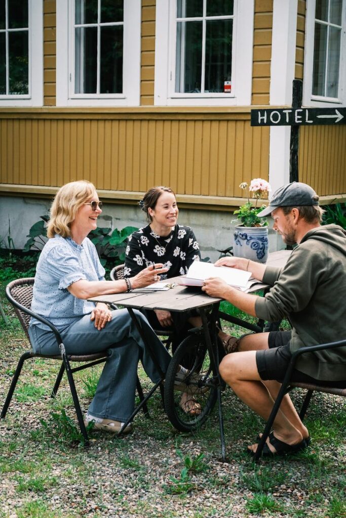 Teamet sitter vid ett utomhusbord och pratar och tittar på papper, med en skylt med texten ”HOTEL” och en gul byggnad i bakgrunden.