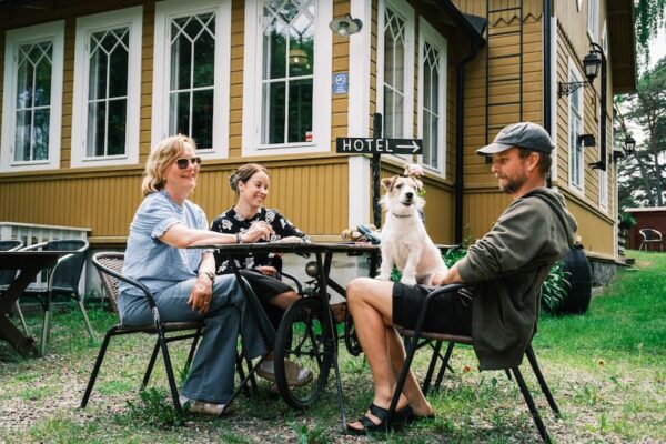 Tre personer och en hund sitter vid ett utomhusbord nära en gul byggnad med en skylt som pekar mot ett hotell.